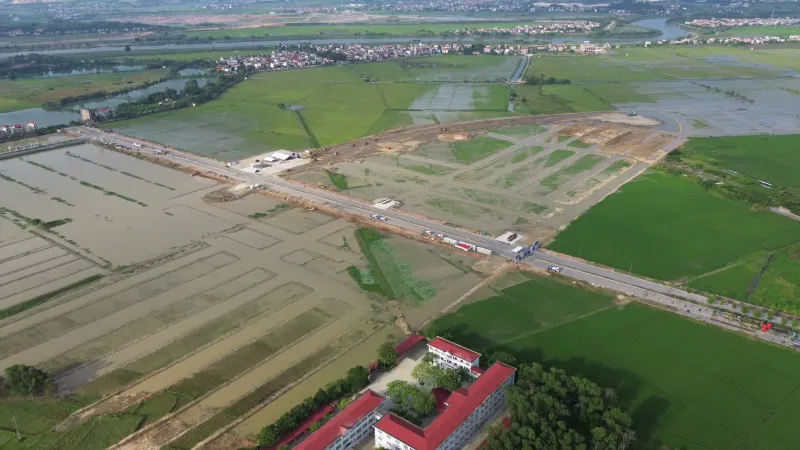 Toàn cảnh công trường từ trên cao, trục đường lớn chạy ngang, phía xa là làng xóm và sông